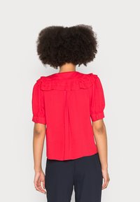 Blusa rossa a maniche corte con scollo arricciato, tessuto testurizzato e maniche con bottoni. Il design sul retro include una piega verticale.