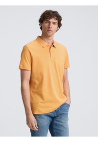 Kortärmad polo skjorta i orange bomull, med krage och tre knappar. Bärs med ljusblå denimjeans.