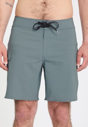 Homme portant un short de bain gris avec un cordon noir, une poche latérale et un tatouage sur le bras gauche, visible de la taille aux genoux.