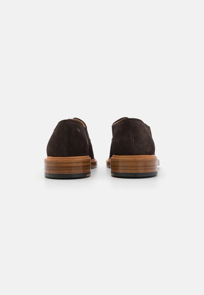 Chaussures en suède marron avec des formes arrondies, dotées d'une semelle en bois lisse avec un bord foncé contrastant. Design épuré et minimal sans matériel visible.