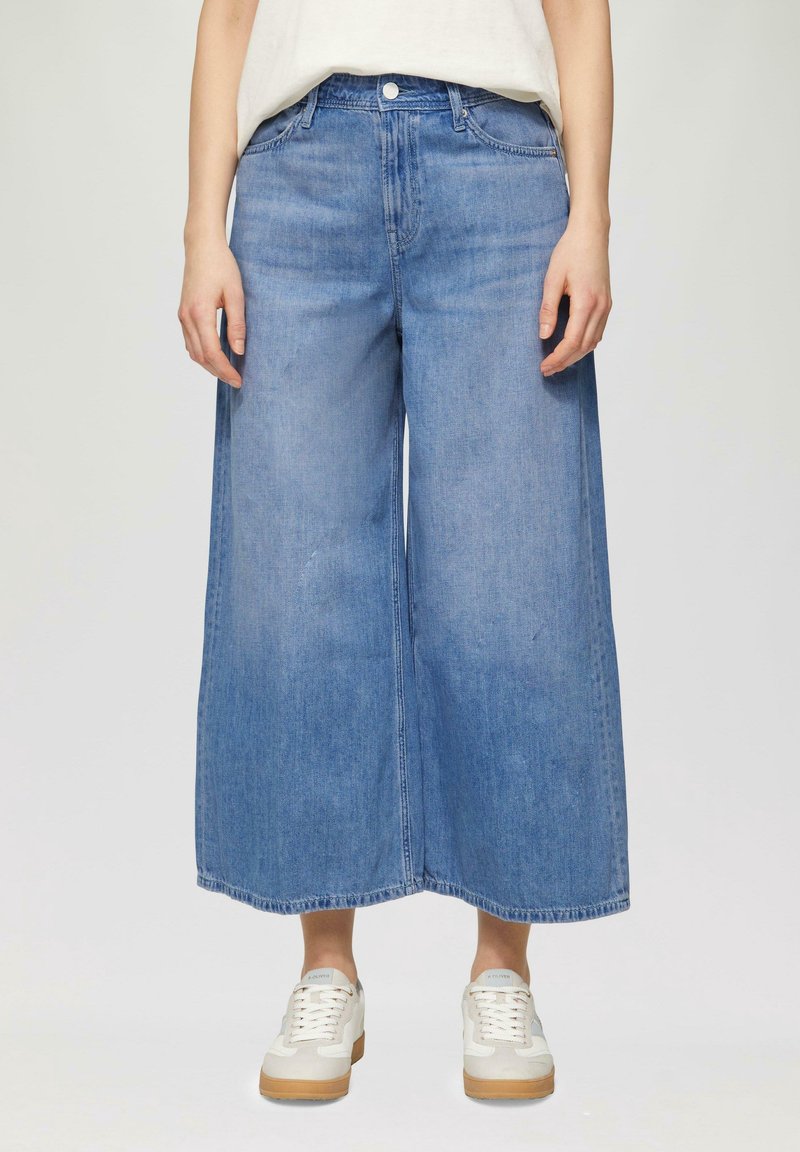 Leveälahkeiset siniset farkkukulotit, joissa on korkea vyötärö, edessä nappikiinnitys ja kaksi sivutaskua, yhdistettynä valkoisiin lenkkareihin.