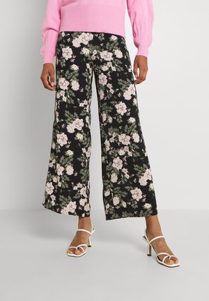 Blumenweite Hosen mit schwarzem Hintergrund, die mit rosa und weißen Blumen bedruckt sind, kombiniert mit weißen Riemchensandalen und einem rosa Pullover.