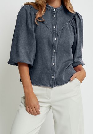 Femme portant un chemisier noir délavé boutonné avec des manches bouffantes et un pantalon blanc taille haute, une main dans la poche.
