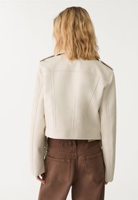 Veste cropped en simili cuir beige avec des épaules structurées, des coutures détaillées et des accents de boutons, associée à un pantalon marron taille haute.