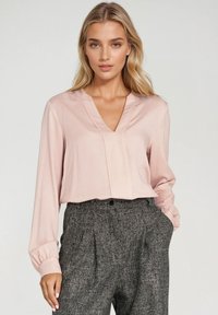 Jasnoróżowa bluzka z dekoltem w serek i długimi rękawami, zdobiona detalami w formie plis. Połączona z szarymi spodniami o wysokim stanie z fakturowanej tkaniny.