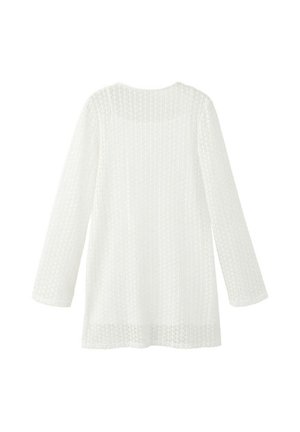 Mujer de pie, vestida con un vestido blanco de encaje de manga larga, sosteniendo un bolso tejido, lleva chanclas negras con tiras beige, mirando hacia la izquierda contra una pared lisa.