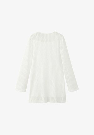 Mujer de pie, vestida con un vestido blanco de encaje de manga larga, sosteniendo un bolso tejido, lleva chanclas negras con tiras beige, mirando hacia la izquierda contra una pared lisa.