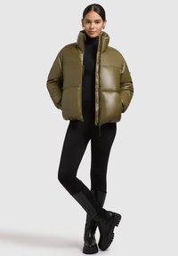Olive green puffer jacket with a high collar, glossy finish, and boxy shape; paired with black leggings and knee-high black boots.