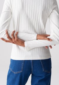 Langärmliges weißes Rippshirt mit strukturierter Stoffoberfläche, eng anliegenden Bündchen und rundem Saum. Kombiniert mit blauen Jeans aus Denim mit Gesäßtaschen.
