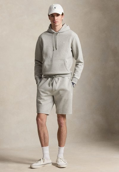 Jeune homme portant un sweat à capuche gris clair, un short assorti, une casquette blanche, des chaussettes et des baskets, debout les mains dans les poches sur un fond neutre.