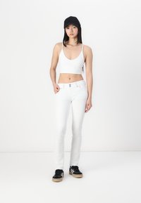 White cropped top, white skinny jeans, and black sneakers with white stripes. Model wears a black cap and stands against a white background.