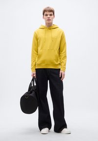 Κίτρινο φούτερ με κουκούλα, μπροστινή τσέπη και κορδόνια, συνδυασμένο με φαρδιά σκούρα τζιν και λευκά αθλητικά παπούτσια, κρατώντας μια μαύρη στρογγυλή τσάντα.