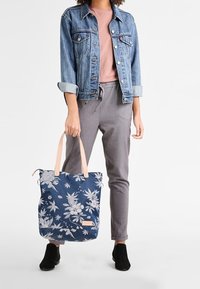 Person wearing denim jacket, pink shirt, gray pants, and black shoes, holding a navy blue tote bag with white floral pattern and beige handles.