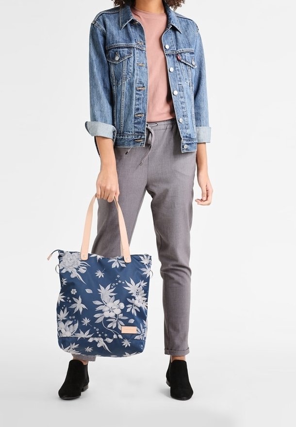 Person wearing denim jacket, pink shirt, gray pants, and black shoes, holding a navy blue tote bag with white floral pattern and beige handles.