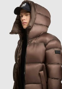 Hnědá péřová bunda s vysokým límcem a oversized kapucí. Obsahuje prošívaný design, lesklou texturu a zapínání na zip. Černá nášivka s logem na rukávu.