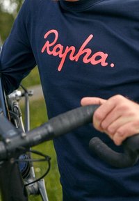Felpa navy con logo "Rapha." rosa, in cotone, colletto a coste, indossata da una persona che tiene il manubrio di una bicicletta.