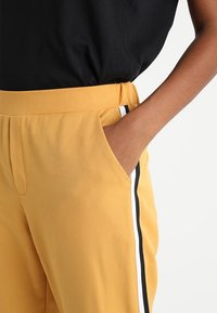 Gula byxor med en slät textur, elastisk midja, sidofickor och svarta och vita ränder längs sidorna.