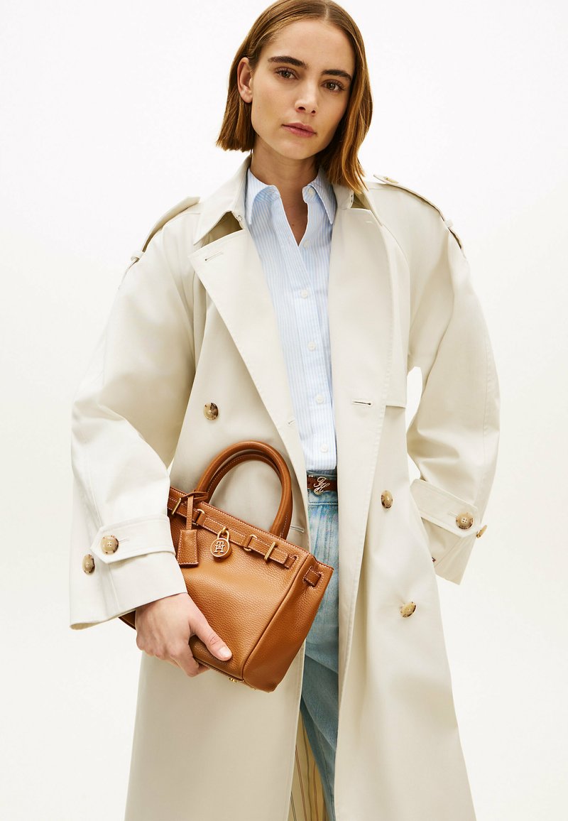 Vrouw met een beige trenchcoat en een lichtblauwe blouse, die een kleine bruine leren handtas met gouden details vasthoudt.
