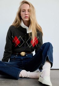 Jeune femme aux longs cheveux blonds assise en tailleur portant un pull à losanges foncé, un jean bleu, des chaussettes blanches et des chaussures blanches à enfiler.