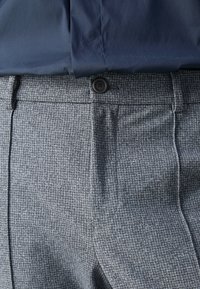 Detailní záběr na kalhoty s šedým houndstooth vzorem s černým knoflíkem, ve kterých je zasunutá tmavě modrá košile.