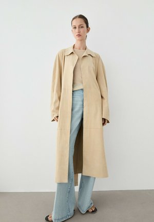 Femme portant un long manteau beige sur un haut beige clair et un jean large bleu clair, debout contre un mur blanc uni.