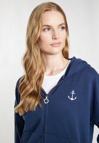 Felpa zip-hoodie blu navy realizzata in tessuto morbido, con un piccolo logo di ancora bianco sul petto e una tasca frontale. Progettata con un cappuccio.