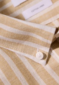 Katoenen-linnen shirt met een lichtgele en witte gestreepte patroon. Heeft een witte knoop en zichtbare stiksels langs de rand van de kraag.