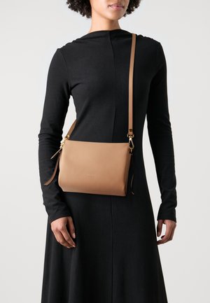 Kožená crossbody taška v hnědé barvě s texturovaným povrchem, nastavitelným popruhem a zlatými kovovými doplňky. Prezentována na pozadí černé šaty.