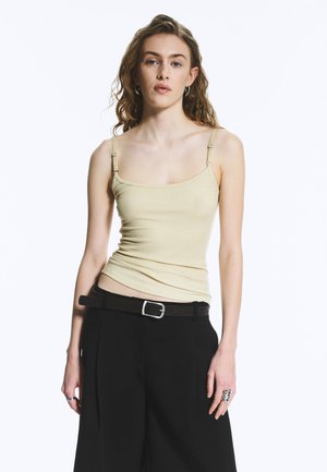 Jeune femme aux cheveux bouclés lâchés, portant un caraco jaune pâle et un pantalon noir taille haute à jambes larges avec une ceinture noire.