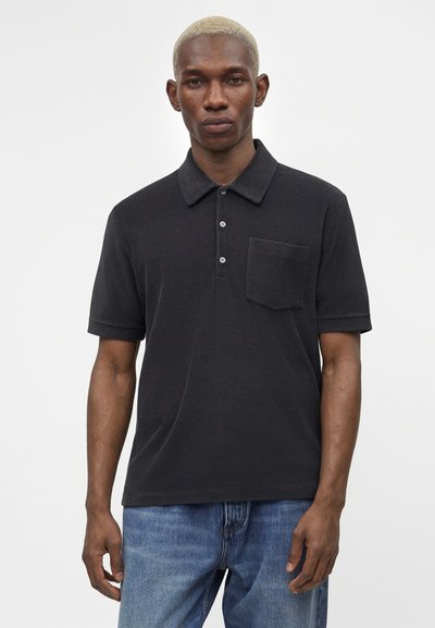 Άνδρας με κοντά ξανθά μαλλιά που φοράει μαύρο κοντομάνικο πόλο με τσέπη στο στήθος και μπλε τζιν, στέκεται μπροστά σε απλό λευκό φόντο.