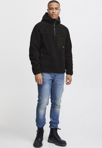 Svart fleece-pullover med hette, med en glidelås lomme på brystet og glidelåser på sidene. Kombinert med lyseblå jeans og svarte snøreboots.