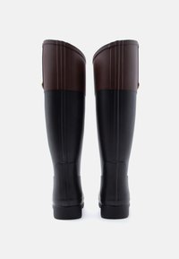 Ψηλές μαύρες λαστιχένιες μπότες με σκούρα καφέ πάνω μέρη, εμφανιζόμενες από πίσω, όρθιες πάνω σε λευκό φόντο.