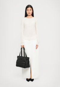 Woman in long white skirt and beige long-sleeve top holding black handbag, wearing black pointed shoes, standing against plain white background.