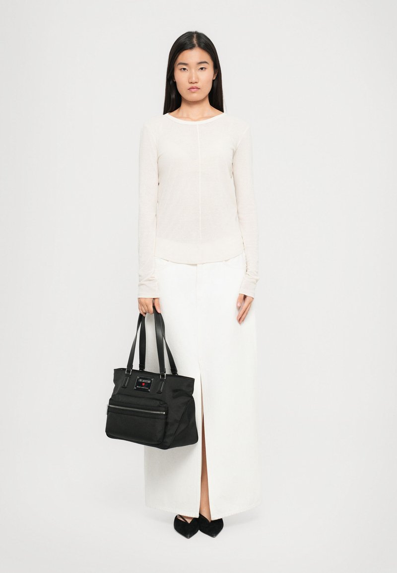 Woman in long white skirt and beige long-sleeve top holding black handbag, wearing black pointed shoes, standing against plain white background.