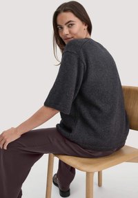 Maglione corto a maniche corte grigio scuro con scollo rotondo e vestibilità ampia. Tessuto a maglia testurizzato. Seduto su una sedia di legno, indossa pantaloni scuri.