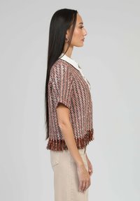 Femme avec de longs cheveux raides portant un haut texturé à manches courtes multicolore avec des franges au bas et un pantalon clair, se tenant de profil.