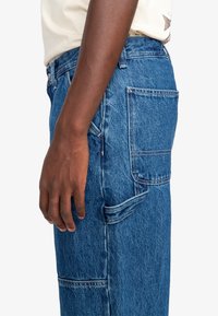 Pantaloni cargo in denim blu con molteplici tasche, texture sbiadita e taglio dritto, dotati di un design a pannello laterale visibile.