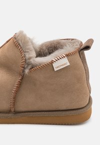 Brune semsket tøffel med myk fleecefôr, kontrasterende sømkanter og en liten stofflapp merket "SHEPHERD" på baksiden.