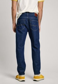 Temno modri jeans z ravnim krojem, dvema zadnjima žepoma in rjavim usnjenim obližem na pasu. Nosi se z rumenimi čevlji.