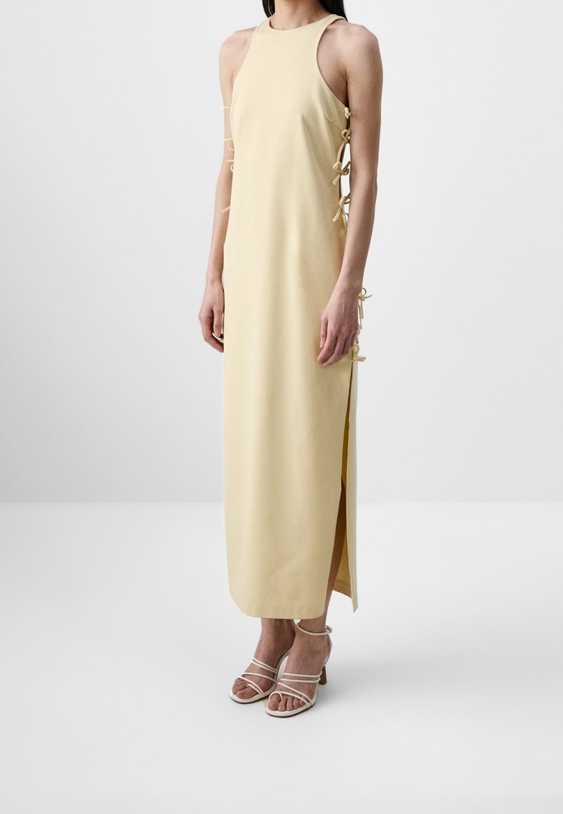 Femme portant une robe maxi jaune pâle sans manches avec des liens et des fentes sur les côtés, assortie à des sandales blanches à talons et à brides.