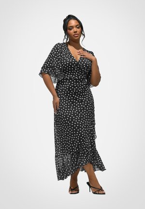 Kvinde iført sort og hvid polkaprikket midikjole med flagrende ærmer og flæsekant, iført sorte remsandaler med høj hæl, poserer mod hvid baggrund.