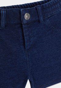 Pantalon en denim bleu foncé avec taille élastique, bouton métallique, passants de ceinture et détail de couture sur la poche avant.