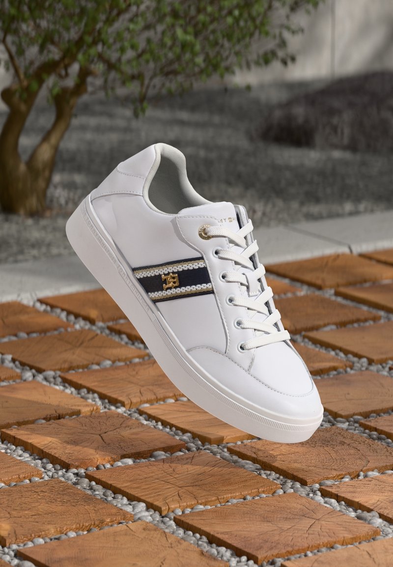 Sneakers bianchi con tomaia in pelle liscia, suola intermedia testurizzata e una striscia blu navy con decorazioni metalliche. Lacci di un bianco sporco.