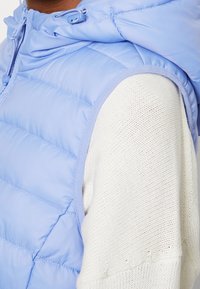 Jasnoniebieska kamizelka puchowa z kapturem, z poziomymi przeszyciami i miękkim białym dzianinowym kontrastem wewnątrz.