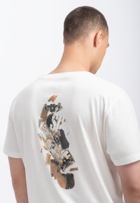 Witte katoenen t-shirt met een grote, abstracte grafische print op de achterkant, waarin tinten bruin, zwart en crème zijn gecombineerd, met skateboard-elementen.