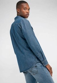 Camicia in denim di blu medio, con chiusura a bottoni, maniche lunghe e spalla a yoke. Abbinata a jeans blu coordinati.