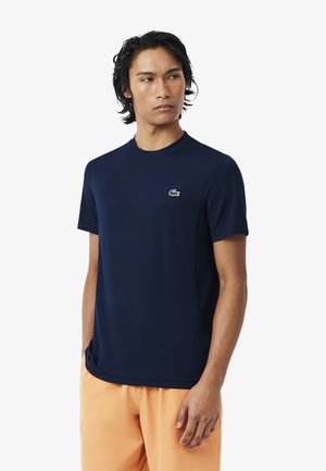 Mann trägt ein marineblaues T-Shirt mit kurzen Ärmeln und grünem Krokodil-Logo sowie hellorangefarbene Hosen, schaut zur Seite.