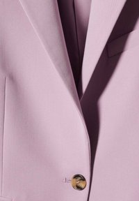 Veste rose clair avec un revers cranté, un bouton doré unique et une texture de tissu lisse, avec un détail de poche sur le côté gauche.