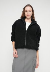 Μαύρο φλις μπουφάν με φερμουάρ, ψηλό γιακά, άνετη εφαρμογή και υφή με ανάγλυφη επιφάνεια. Το μοντέλο φοράει άσπρο μπλουζάκι και γκρι φούστα.