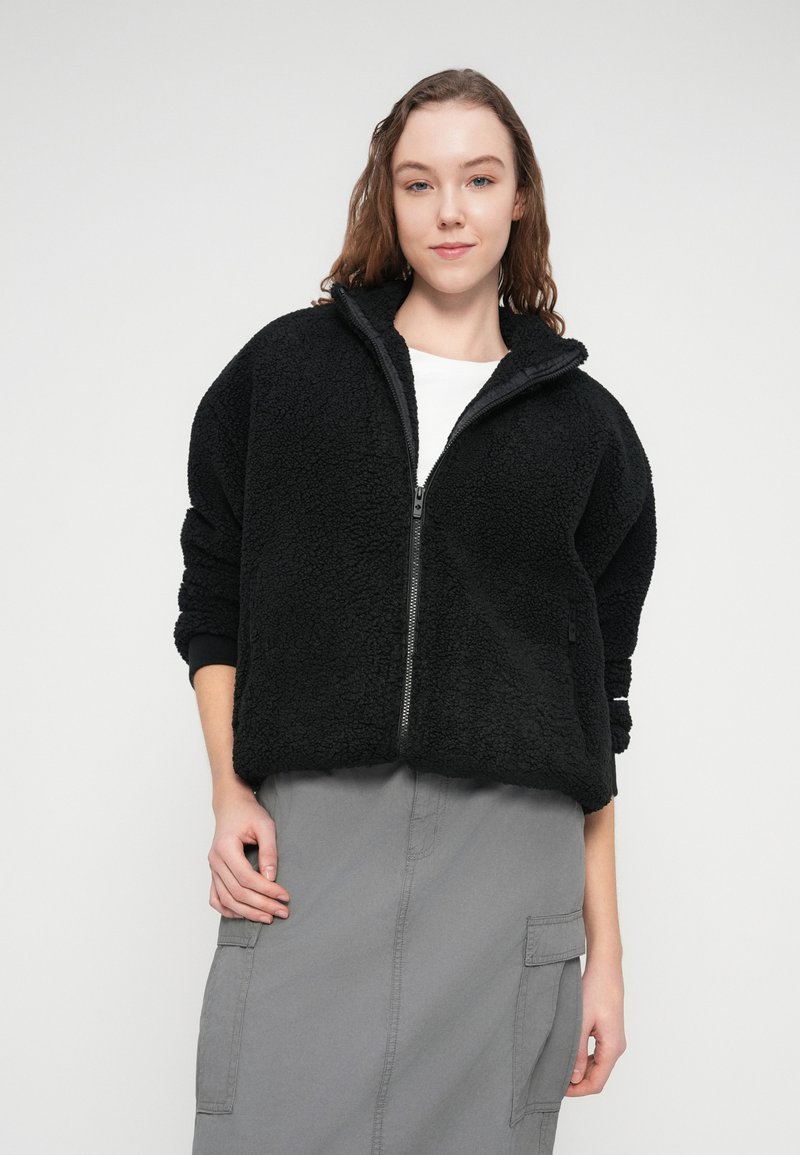 Sort fleecejakke med lynlås, høj krave, løs pasform og tekstureret overflade. Modellen bærer en hvid t-shirt og en grå nederdel.